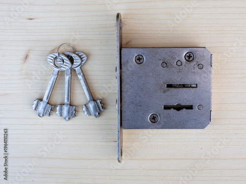 Close-up of an old metal door lock mechanism and a set of vintage keys placed on a light wooden surface. The image highlights classic security hardware, craftsmanship, and retro design