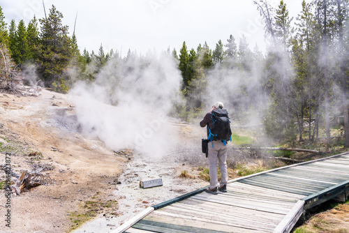 A traveler with a backpack standing on a wooden boardwalk, using a camera to capture the constant steam and mist of Geyser in the Norris Geyser Basin of Yellowstone National Park