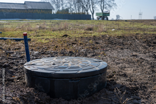 Plastic septic tank cover installed in ground on rural construction site with blue utility pipe and farm buildings in background.