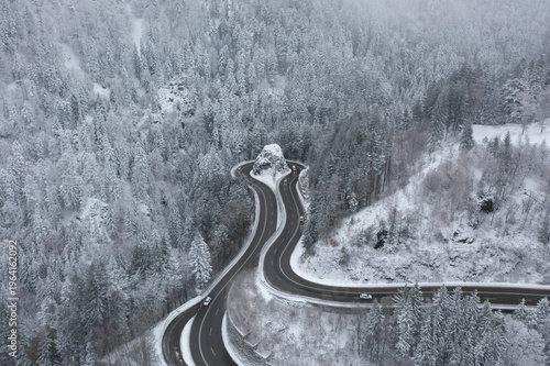 Aerial drone view of winding mountain road curving around rock formation in snowy forest landscape with cars. Scenic winter transportation in Black Forest, Germany