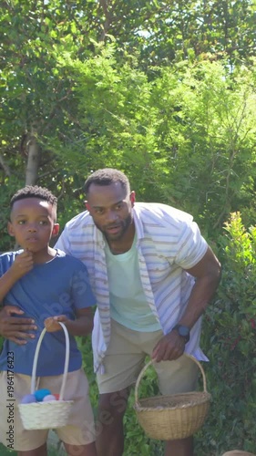 Vertical video: Pointing dad guiding son in blue tee along garden hedge, collecting eggs in baskets