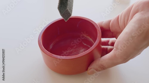 Hand Holding Red Bowl While Applying Paint With Foam Brush in Closeup Crafting Scene