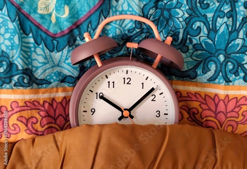 Fotografie Photo of a retro twin-bell alarm clock tucked under a blanket on colorful patterned bedding, morning wake-up concept with cozy home atmosphere and vibrant textile background