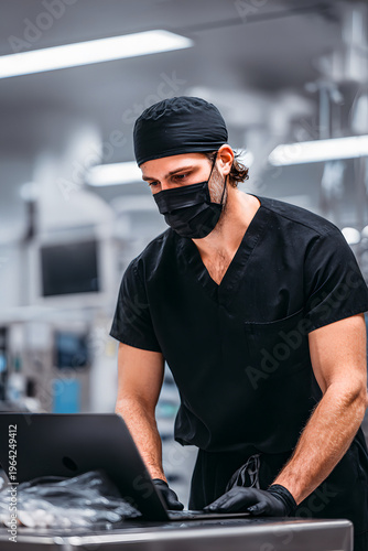 A dental surgeon in a black uniform and face mask carefully reviews a patient's records on a laptop