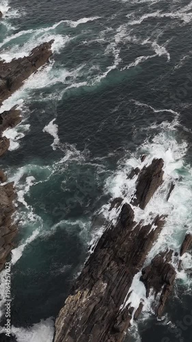 Overhead perspective of dark ocean waves with white foam breaking against a rugged, rocky coastline.