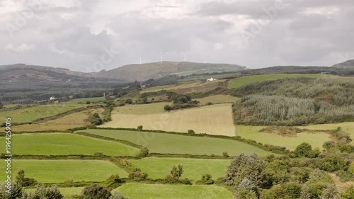 Rolling green hills and patchwork agricultural fields stretch across the rural Irish countryside on a cloudy day.