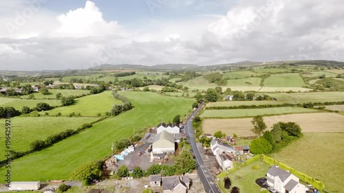 Aerial perspective of a country road running through extensive green fields and scattered homes in the Irish countryside. The daytime landscape is bright under a partially cloudy sky.