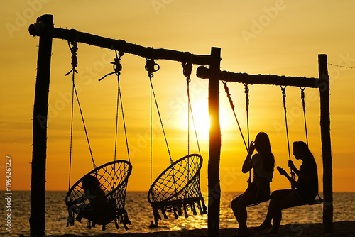 Fotografi Two people sit on swings at a beach as the sun sets behind them