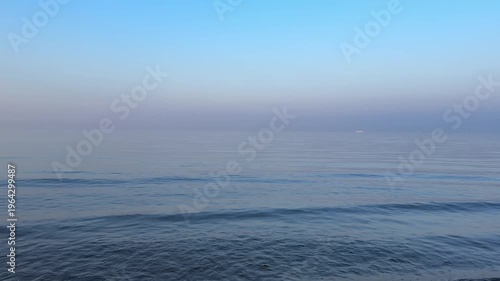 perfectly calm, glassy sea beneath a vast pale blue sky with wispy clouds, where the horizon dissolves into soft coastal mist, creating an exceptionally minimal and serene seascape
