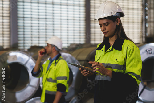 A woman worker wearing a yellow and blue safety vest is holding a tablet in a factory