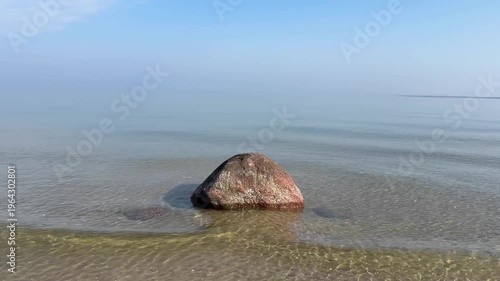 single large dark granite erratic boulder sits half-submerged in a glassy calm sea, perfectly centered against a pale blue hazy horizon, creating a minimalist and meditative coastal composition