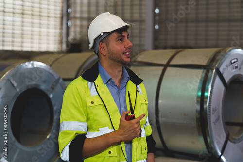 A man in a yellow safety vest is smiling and holding a walkie talkie at warehouse metal sheet.