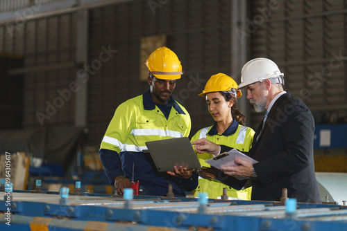 The workers are standing in a factory, looking at a tablet. The engineer talks with the woman's workers. They wear a yellow and white safety helmet for safety.
