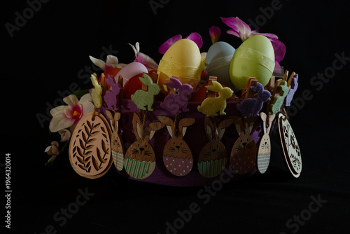 A detailed Easter still life featuring a purple felt basket decorated with a collection of colorful felt and wooden bunny clothespins, filled with various Easter eggs