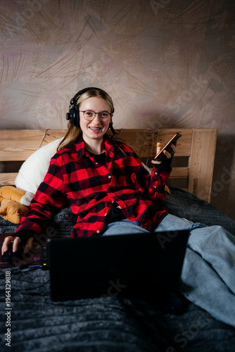 Smiling young woman with braces and glasses wearing headphones sitting on bed with laptop and smartphone at home, remote work or study concept, cozy atmosphere, looking at camera