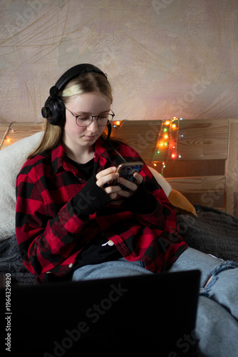 Close-up of a focused young woman with glasses and headphones looking at a smartphone screen, sitting on a bed with blurred Christmas lights in the background, remote work or study from home, cozy