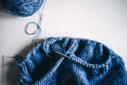 Close-up of metal knitting needles crossed in blue wool fabric, detailed texture of knitwear, darker background, creating handmade clothes concept