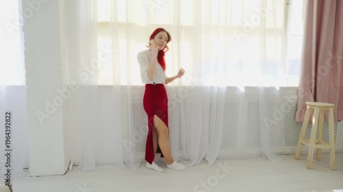 Elegant Young Woman with Red Hair Talking on Smartphone Standing by Large Window with White Curtains in Bright Minimalist Interior