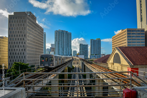 Modern elevated commuter train traveling on tracks through an urban downtown district with high-rise office buildings.