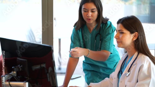 Two female medical professionals, a doctor and an assistant, are in a bright and modern hospital setting, intently reviewing patient results on a computer screen. 