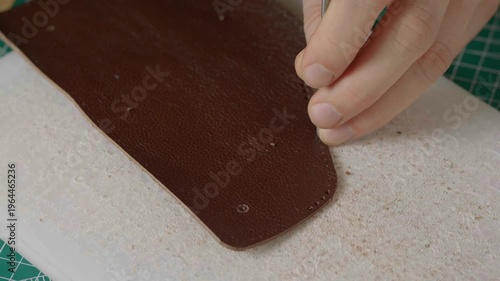 A close-up cinematic shot showing a leather craftsman using a small metal hammer and a round punch to create a series of perfect holes in a strip of natural brown leather.