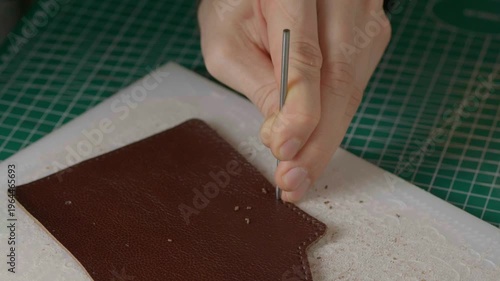 A close-up cinematic shot showing a leather craftsman using a small metal hammer and a round punch to create a series of perfect holes in a strip of natural brown leather.