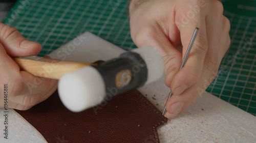 A close-up cinematic shot showing a leather craftsman using a small metal hammer and a round punch to create a series of perfect holes in a strip of natural brown leather.