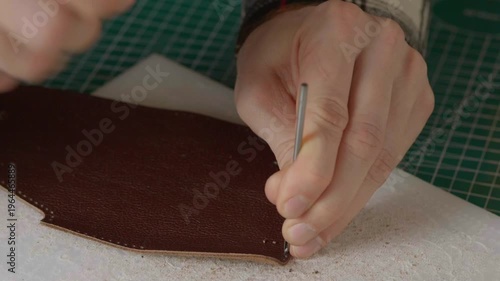 A close-up cinematic shot showing a leather craftsman using a small metal hammer and a round punch to create a series of perfect holes in a strip of natural brown leather.