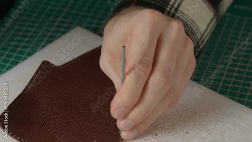 A close-up cinematic shot showing a leather craftsman using a small metal hammer and a round punch to create a series of perfect holes in a strip of natural brown leather.