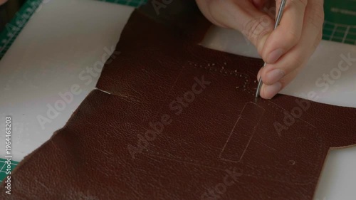 A close-up cinematic shot showing a leather craftsman using a small metal hammer and a round punch to create a series of perfect holes in a strip of natural brown leather.