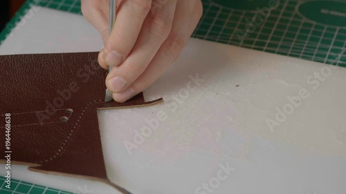 A close-up cinematic shot showing a leather craftsman using a small metal hammer and a round punch to create a series of perfect holes in a strip of natural brown leather.