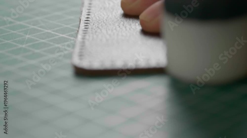 A very detailed close-up of a craftsman crafting a genuine brown leather item, perhaps a wallet or key holder. He's just glued two pieces together and is pressing them together with small hammer.