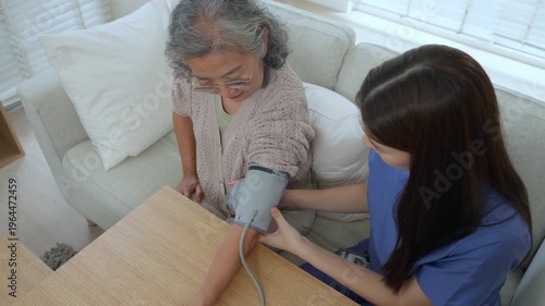 Caregiver elderly care home nurse asian senior woman checks blood pressure with cuff during calm visit, support at table in bright living room, health monitor on wood surface, trust and focus