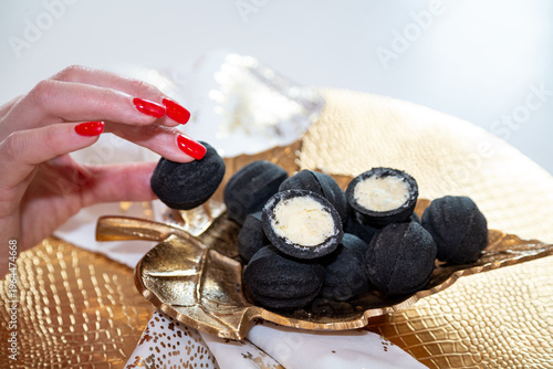 Black nut-shaped cookies with filling on a golden leaf-shaped plate on a table in the rays of the sun and a girl's fingers with a red manicure