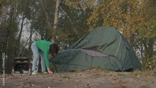 Caucasian woman erecting green tent camping in autumn forest, concentrating on frame assembly and securing flysheet parked car and birch trees in background, fallen leaves underfoot, overcast soft