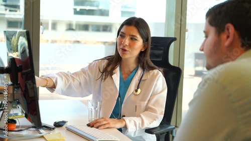 A female doctor in a white coat sits at a desk with a computer, explaining test results to a male patient in a modern office setting with natural light.