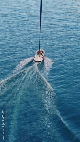 Aerial view of a speedboat pulling a parasail over calm sea, leaving wake trails. Scenic coastline with mountains and city in soft evening light, travel and adventure concept.