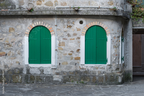 Historic stone wall with arched windows and green wooden shutters in Grado, Italy. Rustic Mediterranean architecture in the old town alley, traditional Italian building facade with brick details.