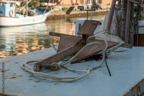 Close-up of a rusty iron anchor with white rope on a fishing boat in Grado, Italy. Traditional nautical equipment in the canal port harbor with blurred water and vessels in the background.