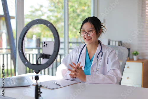 Young female doctor wearing a lab coat and stethoscope, smiling while recording a video on a smartphone for remote healthcare consultation