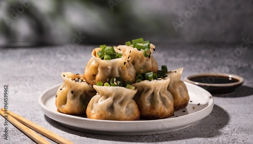 A plate of golden brown pan fried dumplings garnished with chopped green onions and sesame seeds sits on a textured gray surface with a dish of soy sauce and chopsticks