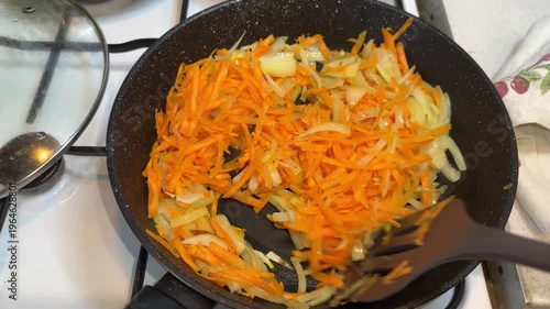 Grated carrots and sliced onions sautéing in a dark frying pan on a gas stove.