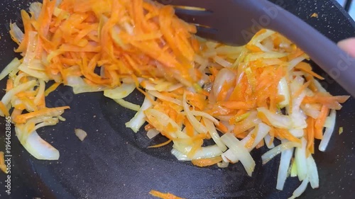 Grated carrots and sliced onions sautéing in a dark frying pan on a gas stove.