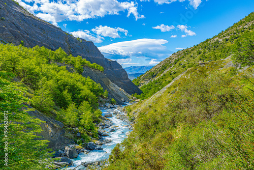 Tablou pe pânză A rushing stream flows through a deep, sunlit valley with steep rocky slopes and lush green vegetation under a blue sky