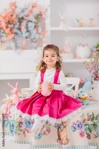 little girl in pink dress holding easter egg while sitting on table with pastel decorations, soft spring interior and cozy festive home mood