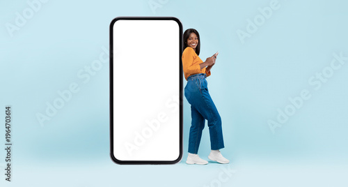 Canvastavla Cheerful young black woman using mobile phone standing near giant smartphone with empty screen on blue studio background, full body length