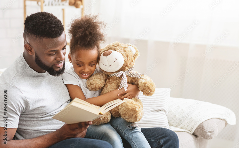 Obraz premium Cute African American family father and daughter reading book, sitting on couch at home, copy space