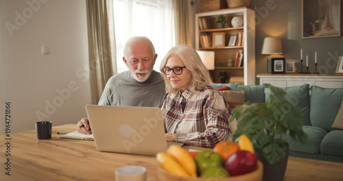 Two elderly individuals sit together at a table with a laptop, reviewing life insurance plans and discussing financial choices in their living room with natural light.