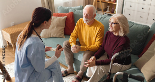 Nurse sits on the floor and talks to an elderly couple on the couch in their living room. The couple listens and engages with the nurse during the home visit.