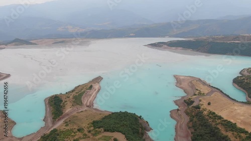Aerial view of an industrial tailings dam in a mountainous region. The reservoir features unnaturally bright turquoise water mixing with a large expanse of grey, sludgy sediment.